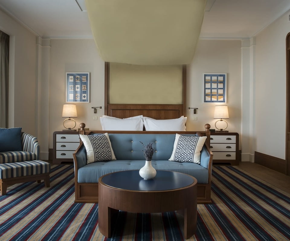 A modern and elegantly decorated hotel room featuring a wooden bed with plush bedding, striped upholstered chairs, a round coffee table, and a colorful striped rug.