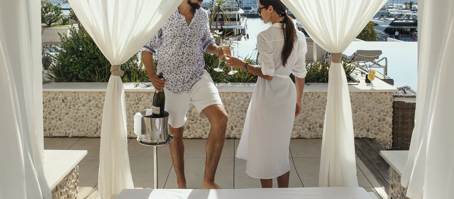 A man in a patterned short-sleeve shirt and white shorts stands next to a metal ice bucket with a bottle, handing a glass of sparkling wine to a woman.