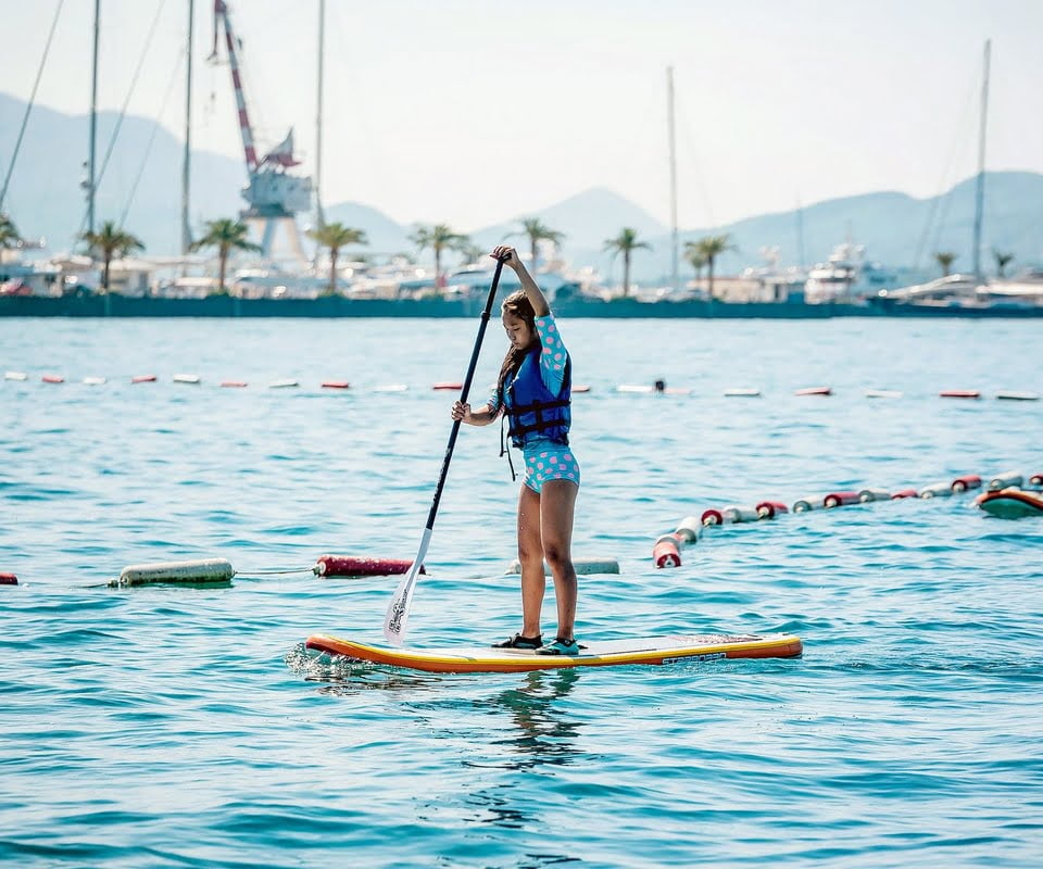 Woman paddleboarding in calm sea waters with a mountainous backdrop and yachts in the distant harbour, under a clear blue sky.