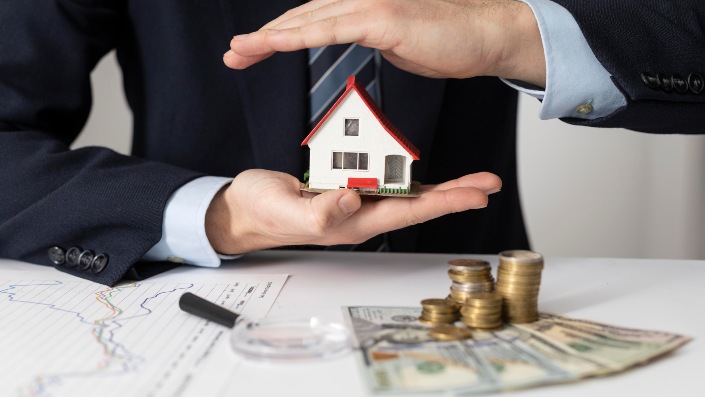 A person in a business suit holding a small model house, with one hand above and one hand below, next to a stack of coins and dollar bills on a table.