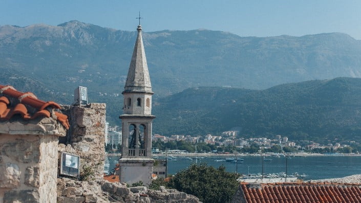 Scenic coastal town with a bell tower, historic architecture, boats along the shore, and mountains in the background.