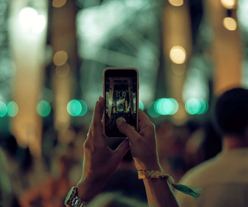 Amidst the electrifying atmosphere of Rubix Festival in Porto Montenegro, an attendee captures the essence of a pulsating concert crowd with their phone. Vibrant green lights illuminate the scene, highlighting the sea of raised hands and the energy of the moment.