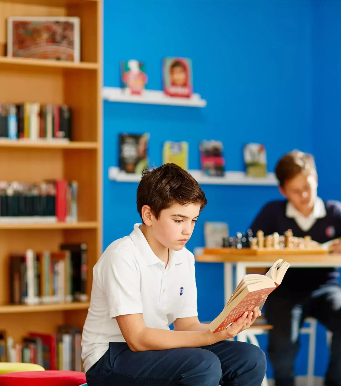 Young boy focused on reading a book in a brightly colored classroom, with another student playing chess in the background, surrounded by books and educational materials.