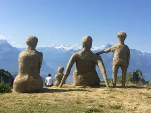Large straw sculptures resembling human figures overlooking a mountain landscape.
