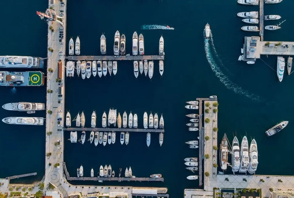 A top-down aerial view of Porto Montenegro Marina, featuring neatly arranged yachts and superyachts docked along the jetties, with boats navigating through the deep blue waters.