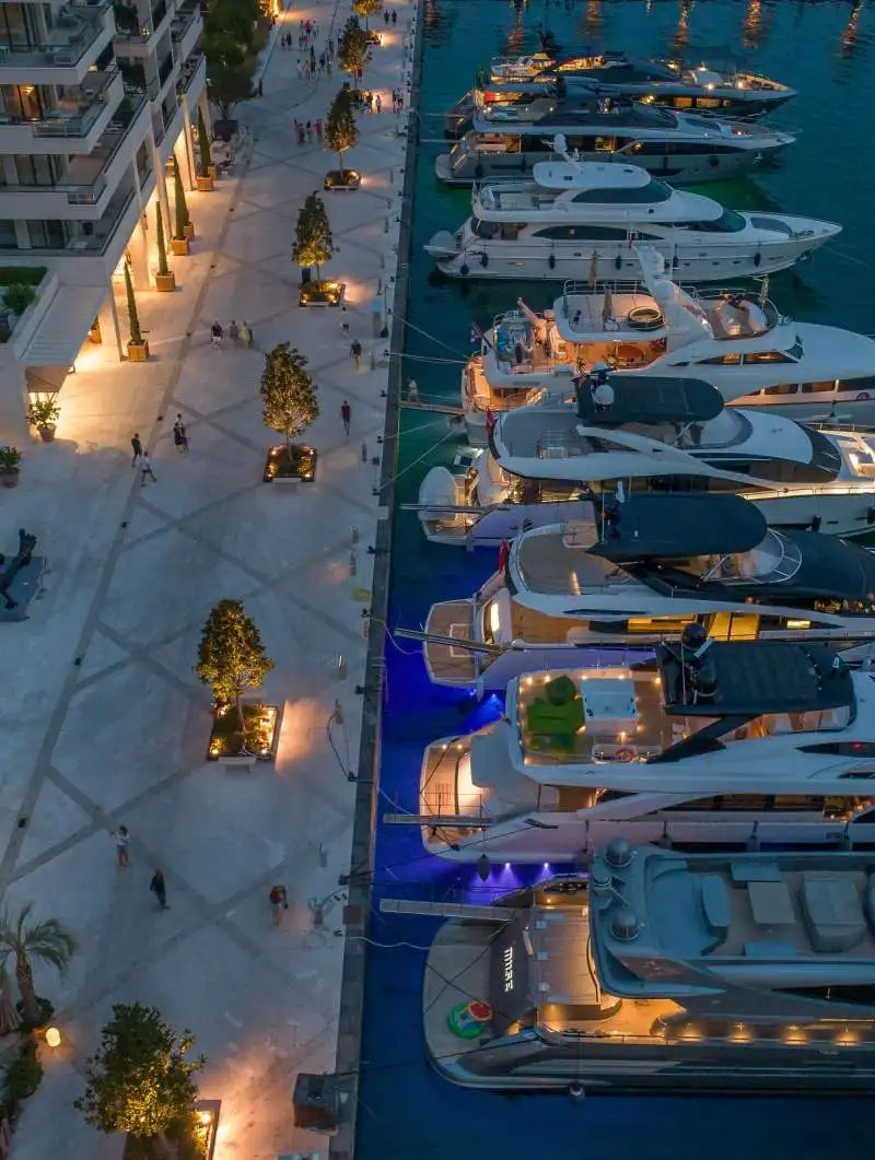Aerial view of a luxury marina at dusk, with yachts illuminated by soft golden lights. A pedestrian promenade lined with trees and warm-lit pathways runs parallel to the moored yachts, creating a vibrant evening atmosphere.