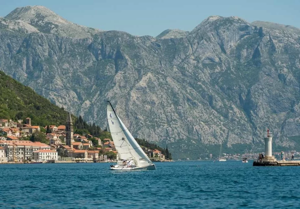 A PMYC sailboat gliding through the Bay of Kotor near the historic town of Perast, Montenegro, with dramatic mountain scenery and a lighthouse in the background.