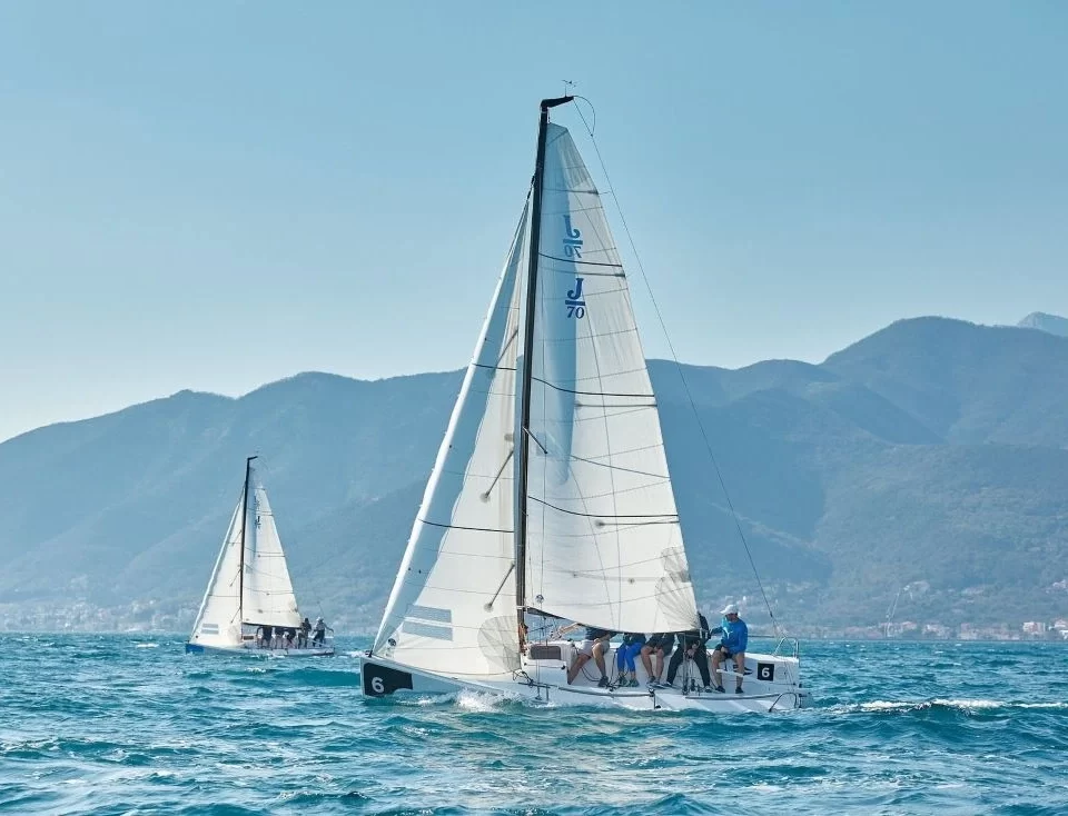 Two sailboats racing in the PMYC Sailing Squadron, gliding across the deep blue waters with a stunning mountainous backdrop under clear skies.