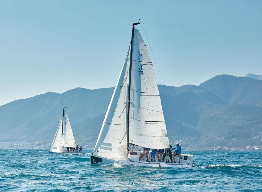 Two sailboats racing in the PMYC Sailing Squadron, gliding across the deep blue waters with a stunning mountainous backdrop under clear skies.