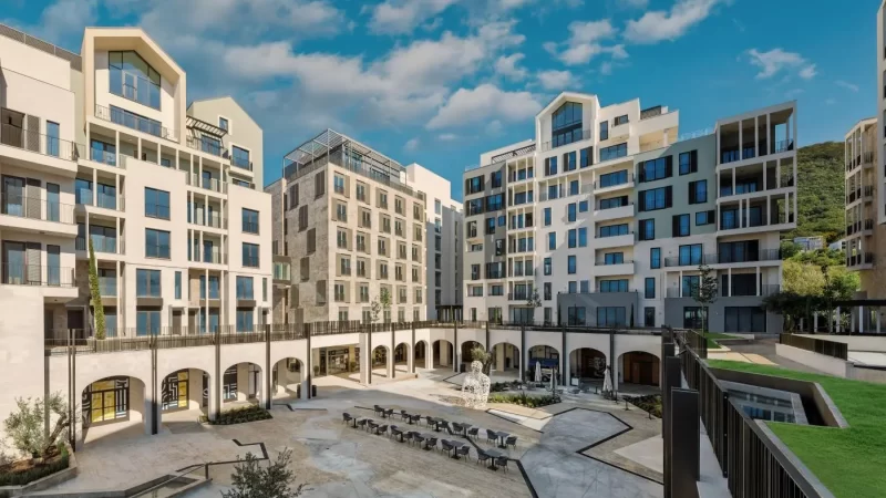 Modern Mediterranean-style residential and commercial buildings surrounding a spacious plaza with outdoor seating, architectural arches, and a centerpiece sculpture, set against a backdrop of greenery and a clear blue sky, Boka Place neighborhood.