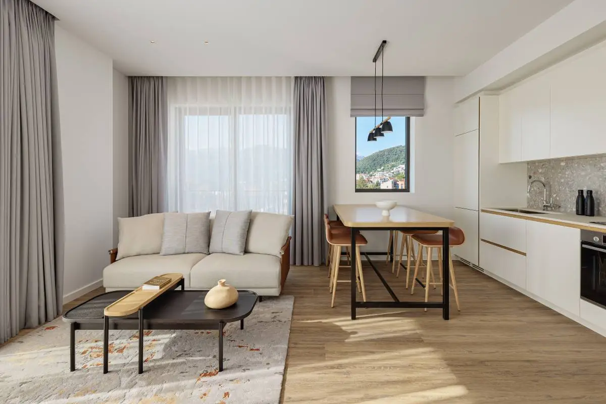 Modern open-concept living room and dining area featuring a neutral-tone sofa, minimalist coffee table, and a wooden dining table with leather chairs. The room is brightened by natural light from large windows with sheer curtains, showcasing a scenic view of greenery.