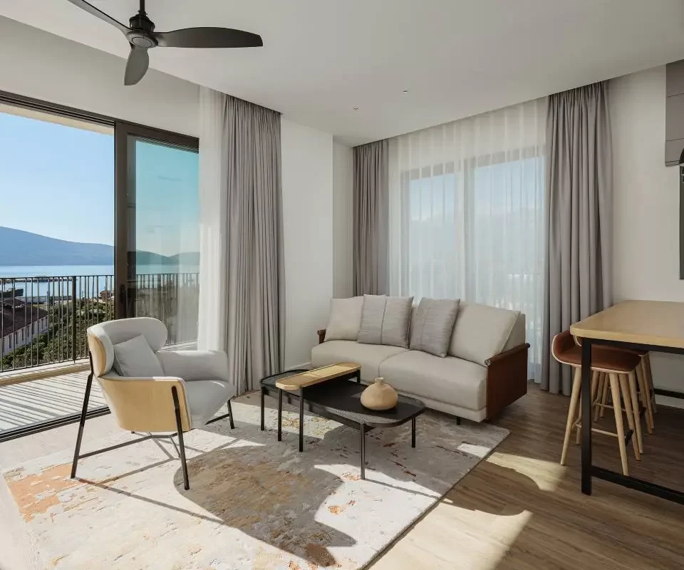 Modern living and dining area in Siro Residences featuring a neutral-tone sofa, sleek armchair, and black coffee table on a textured rug. Large sliding doors open to a balcony with sea views, while a dining table with bar stools sits near a window showcasing mountain scenery.