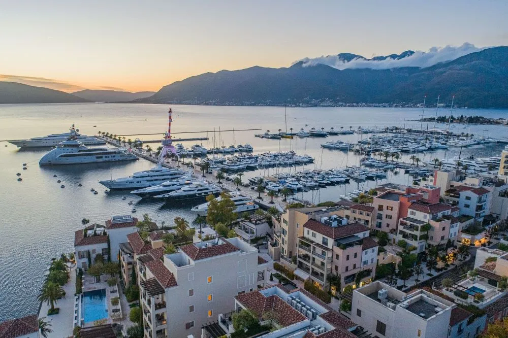 Scenic aerial view of Porto Montenegro Marina at dusk, with superyachts and sailboats moored along the waterfront, surrounded by luxury residences and mountains in the background.