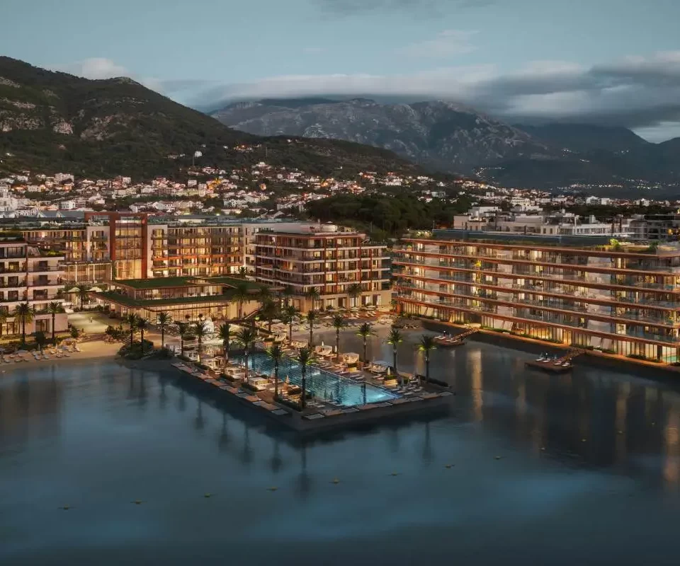 Elegant beachfront Synchro Yards residences at dusk, featuring modern architecture with warm lighting, surrounded by serene waters, lush palm trees, and a marina, all set against the backdrop of majestic mountains and a quaint coastal town.