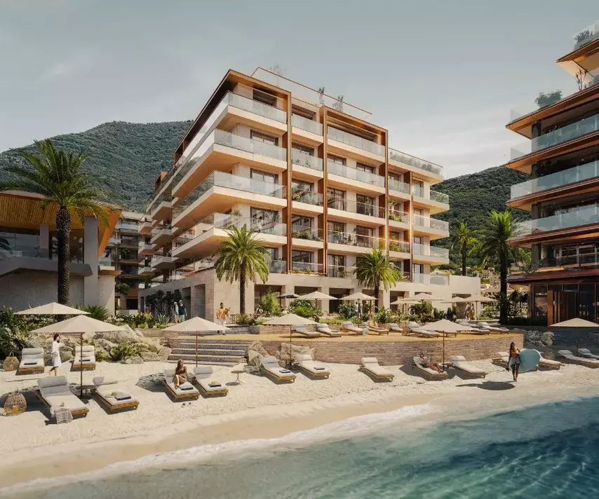 Daytime view of the IVO Residences in Porto Montenegro, a luxurious beachfront apartment complex with multiple balconies, palm trees, and sun loungers on a sandy beach.